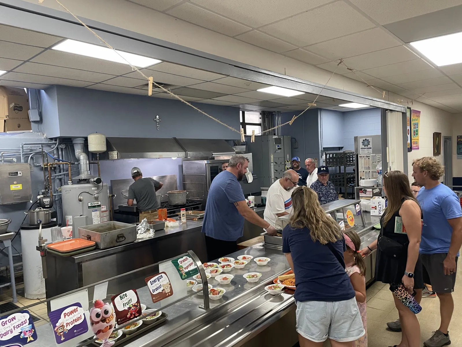 Wide view of the school kitchen with volunteers cooking and families waiting in line