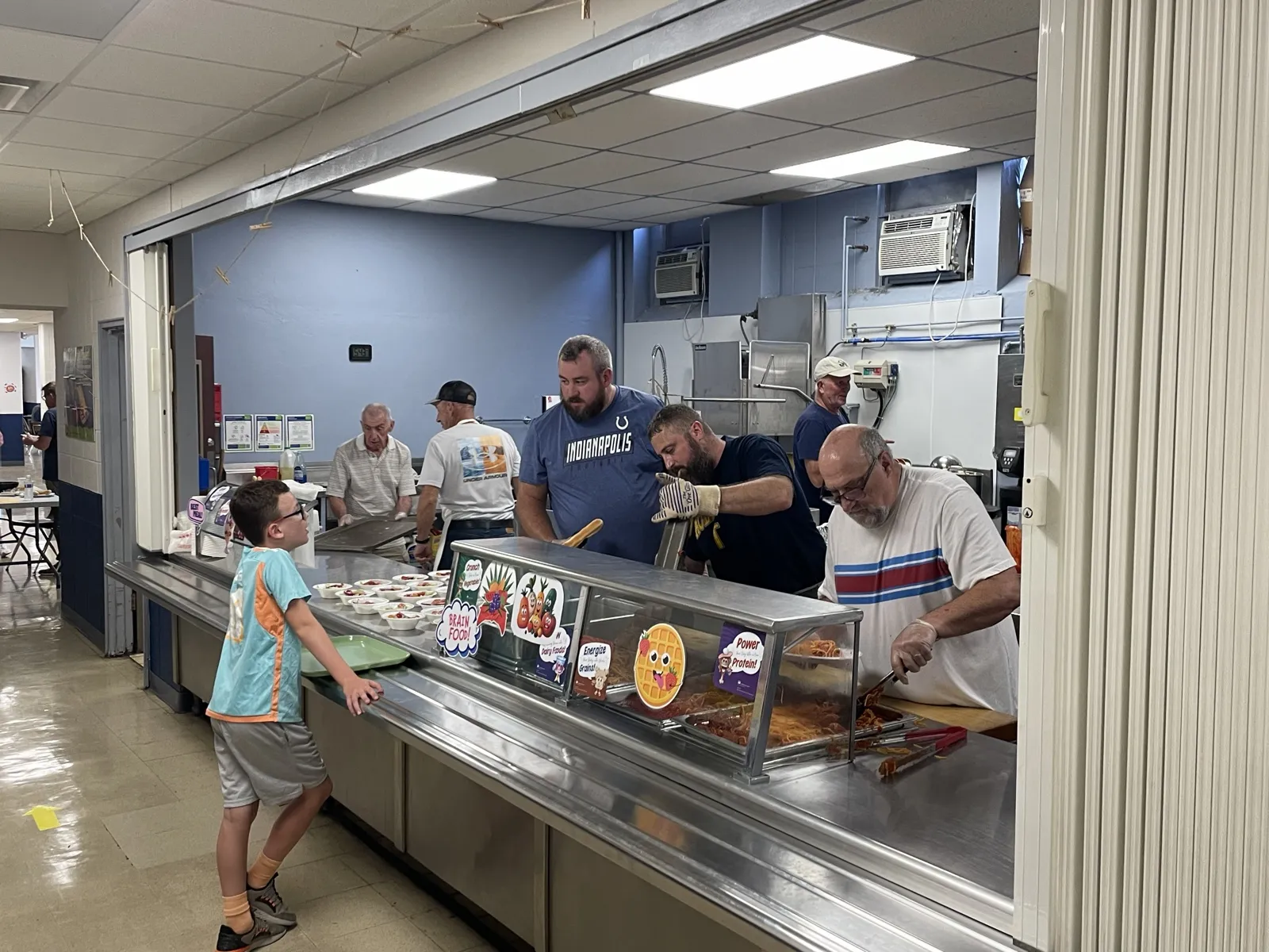 A young guest sliding a tray along the counter as volunteers serve spaghetti and bread